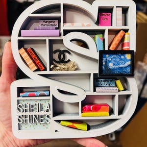 May include: A white, S-shaped shelf with multiple compartments, holding miniature books, a framed painting of Van Gogh's Starry Night, and a Chanel logo. The shelf also features the text "Sheila Shines."