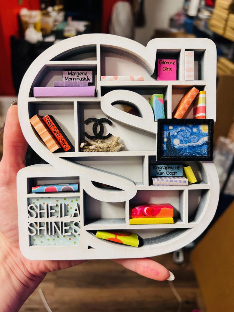 May include: A white, S-shaped shelf with multiple compartments, holding miniature books, a framed painting of Van Gogh's Starry Night, and a Chanel logo. The shelf also features the text "Sheila Shines."
