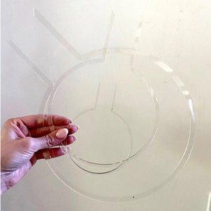 May include: A clear plastic stencil with three concentric circles and three radiating lines. The stencil is held in a hand.