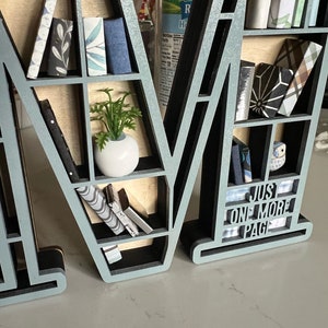 K&ouml;nnte beinhalten: Ein hellblaues Holzregal in Form des Buchstabens "M" mit Regalbrettern, die mit Miniaturb&uuml;chern, Pflanzen und einer kleinen Eulenfigur gef&uuml;llt sind. Das untere Regal tr&auml;gt den Text "Just One More Page".