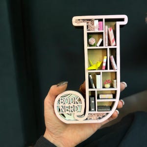 May include: A pink wooden letter J shaped like a bookshelf with miniature books, a flower, and a paper crane inside. The letter has the words "Book Nerd" on the bottom.