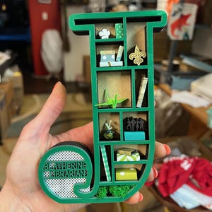 May include: A green wooden shelf in the shape of the letter "J" with miniature decorative items inside. The shelf contains a chef's hat, books, a fleur-de-lis, and a paper crane. The text "BEATHERING LIBRARIAN" is on the circular base.
