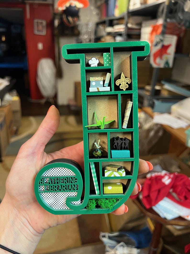 May include: A green wooden shelf in the shape of the letter "J" with miniature decorative items inside. The shelf contains a chef's hat, books, a fleur-de-lis, and a paper crane. The text "BEATHERING LIBRARIAN" is on the circular base.