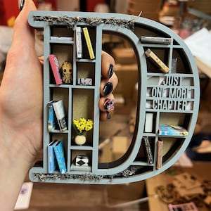 May include: A gray wooden letter "D" shaped bookshelf, filled with miniature books, a Groot figurine, and a small vase of yellow flowers. The text "Just One More Chapter" is visible. The shelf is decorated with moss.