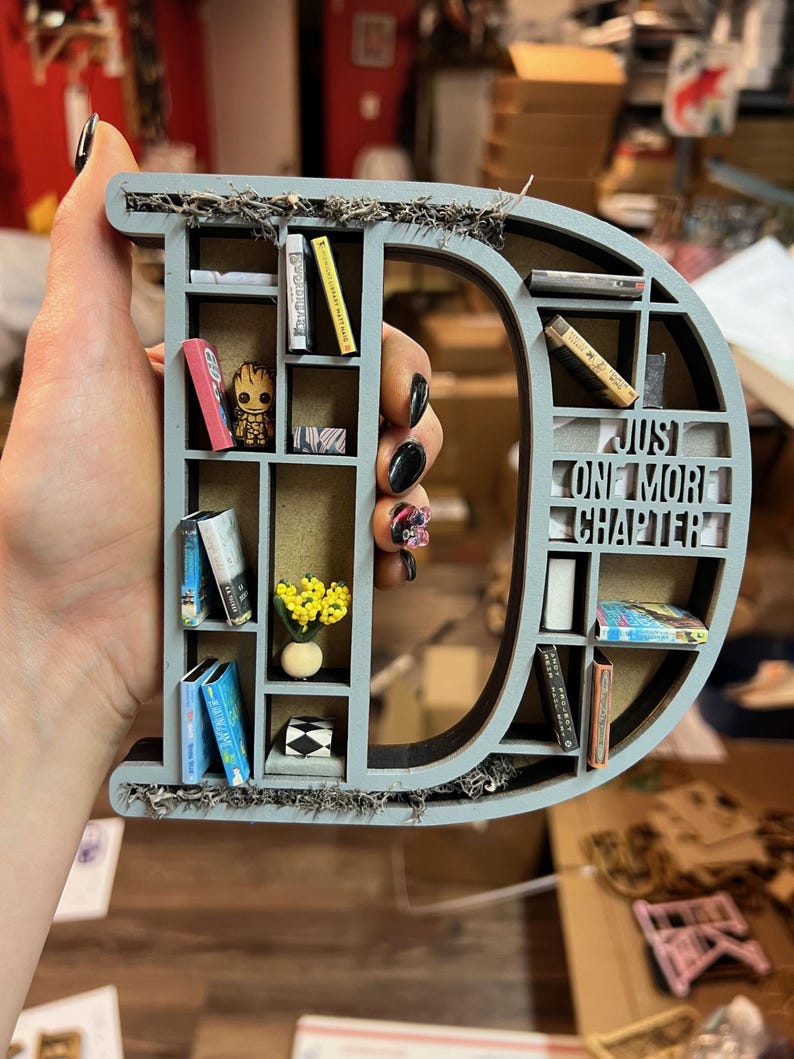 May include: A gray wooden letter "D" shaped bookshelf, filled with miniature books, a Groot figurine, and a small vase of yellow flowers. The text "Just One More Chapter" is visible. The shelf is decorated with moss.