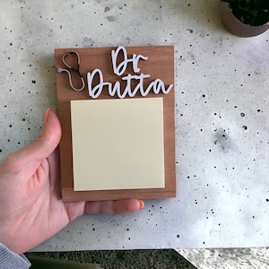 May include: A wooden memo pad with the name "Dr. Dutta" in white script. A small stethoscope is attached to the top left. A yellow sticky note pad is adhered to the wooden base. The memo pad is being held by a person.