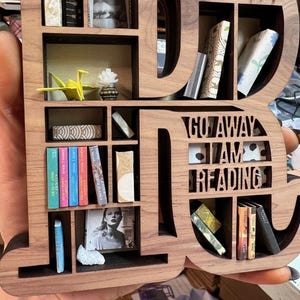 May include: Wooden bookshelf shaped like the word "DI", filled with miniature books, a photo, and decorative items. The text "GO AWAY I AM READING" is cut out of the wood. A unique gift for book lovers.