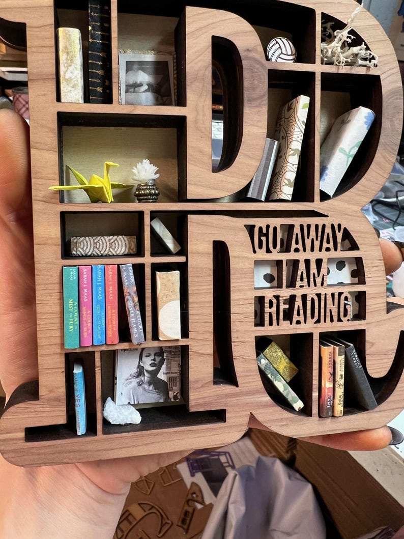 May include: Wooden bookshelf shaped like the word "DI", filled with miniature books, a photo, and decorative items. The text "GO AWAY I AM READING" is cut out of the wood. A unique gift for book lovers.