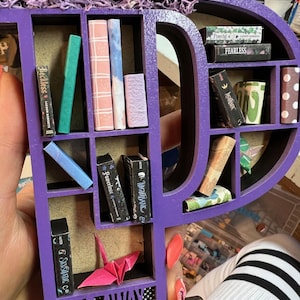 May include: A purple wooden shelf in the shape of the letter "P" filled with miniature books of various colors and sizes. A pink origami crane and the words "GO AWAY I AM READING" are also visible. The shelf is decorated with purple moss.
