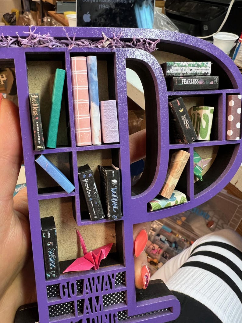 May include: A purple wooden shelf in the shape of the letter "P" filled with miniature books of various colors and sizes. A pink origami crane and the words "GO AWAY I AM READING" are also visible. The shelf is decorated with purple moss.