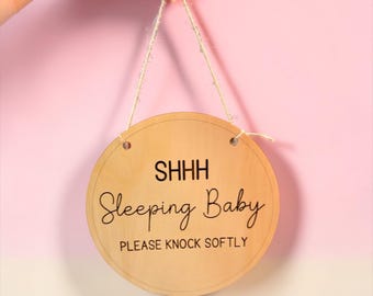 May include: A round wooden sign with the text "SHHH Sleeping Baby PLEASE KNOCK SOFTLY" in black script. The sign is suspended by a natural twine string and is held against a pink background.