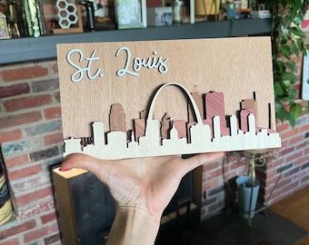 May include: A wooden cutout of the St. Louis skyline, featuring the iconic Gateway Arch. The cutout is painted in shades of brown and red, and the text "St. Louis" is written in white on the top of the cutout.