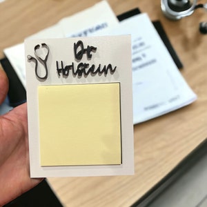 以下が含まれることがあります： 黒い聴診器と黒い筆記体で「Dr. Holstein」と書かれた白い看板。黄色の付箋が看板に貼られている。