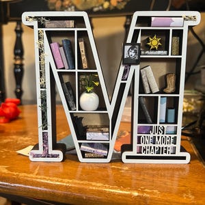 May include: A white and black wooden shelf in the shape of the letter "M" filled with miniature books, a small photo, and decorative items. The text "JUST ONE MORE CHAPTER" is visible on the bottom shelf. The shelf is on a wooden table.