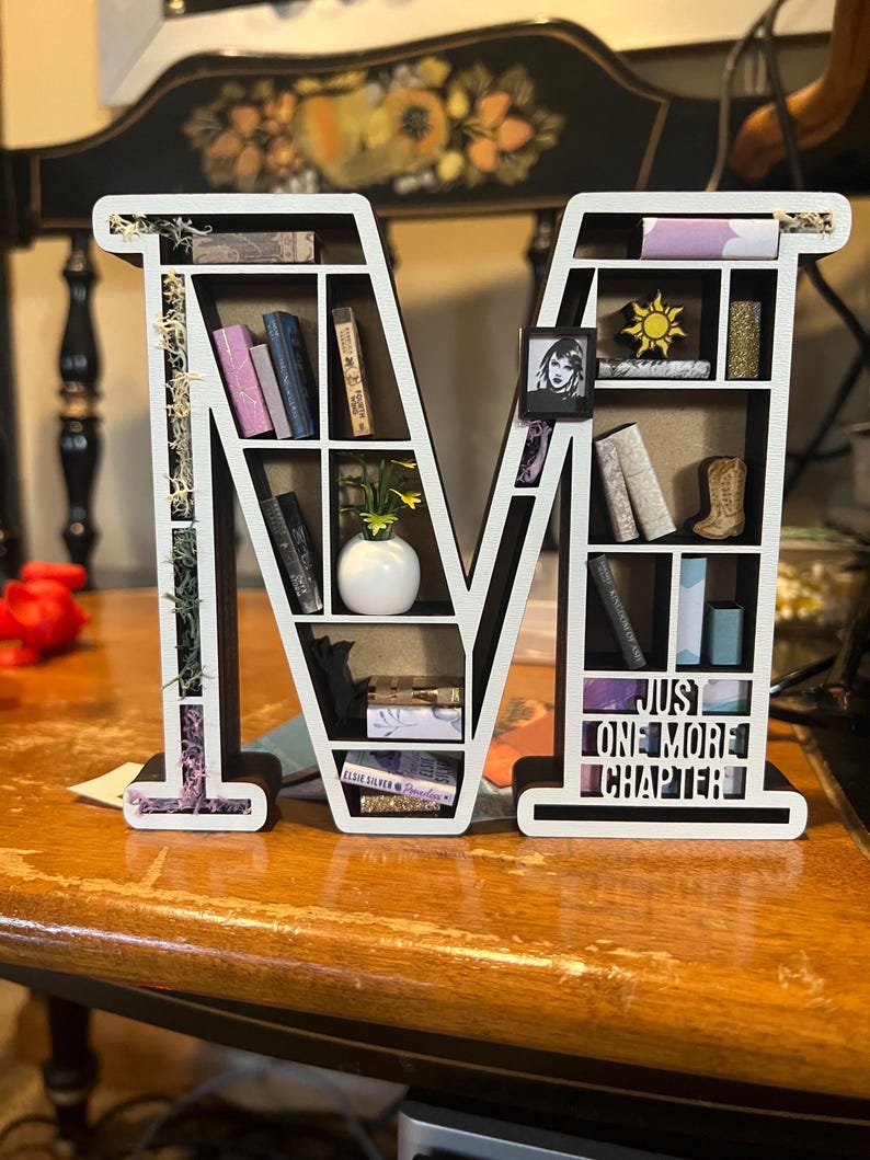May include: A white and black wooden shelf in the shape of the letter "M" filled with miniature books, a small photo, and decorative items. The text "JUST ONE MORE CHAPTER" is visible on the bottom shelf. The shelf is on a wooden table.