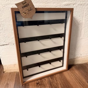 May include: A wooden framed display case with a glass front. The case has multiple horizontal shelves with small hooks. A wooden shield with the text "Bark Ranger Kali" and a dog illustration is attached to the top of the frame.