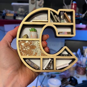 K&ouml;nnte beinhalten: Ein goldfarbenes und schwarzes Regal in Form des Buchstabens C mit mehreren Regalbrettern. Die Regalbretter sind mit Miniaturb&uuml;chern, einer kleinen Topfpflanze und einem goldenen und korkartigen Hintergrund gef&uuml;llt.