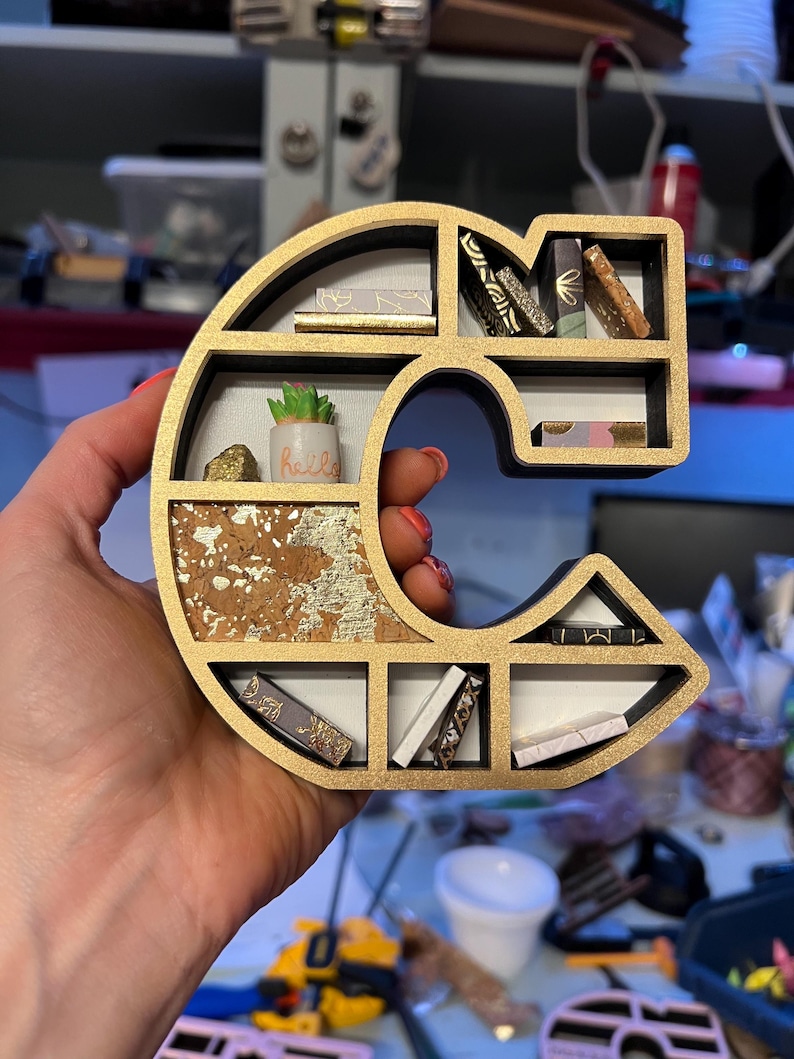 K&ouml;nnte beinhalten: Ein goldfarbenes und schwarzes Regal in Form des Buchstabens C mit mehreren Regalbrettern. Die Regalbretter sind mit Miniaturb&uuml;chern, einer kleinen Topfpflanze und einem goldenen und korkartigen Hintergrund gef&uuml;llt.