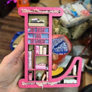 K&ouml;nnte beinhalten: Ein rosa Holzbuchstabe L in Form eines B&uuml;cherregals mit den Worten "Go Away I Am Reading" auf der Vorderseite. Die Regale sind mit Miniaturb&uuml;chern und anderen Dekorationsartikeln gef&uuml;llt.