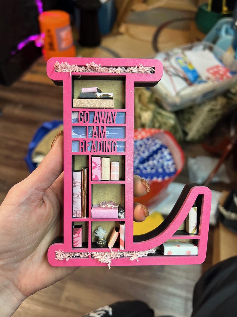 K&ouml;nnte beinhalten: Ein rosa Holzbuchstabe L in Form eines B&uuml;cherregals mit den Worten "Go Away I Am Reading" auf der Vorderseite. Die Regale sind mit Miniaturb&uuml;chern und anderen Dekorationsartikeln gef&uuml;llt.