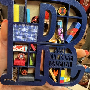 May include: A navy blue wooden shelf in the shape of the letters "DC" with various miniature books and superhero-themed figurines. The shelf includes the text "JUST ONE MORE CHAPTER".