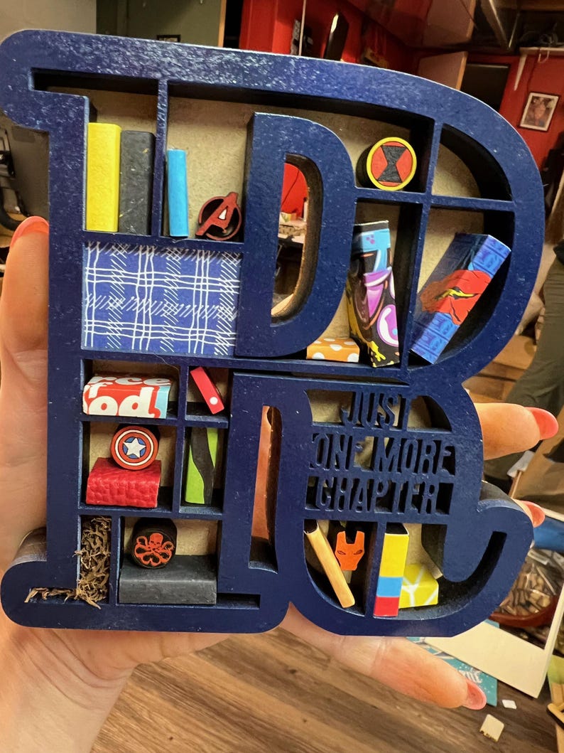 May include: A navy blue wooden shelf in the shape of the letters "DC" with various miniature books and superhero-themed figurines. The shelf includes the text "JUST ONE MORE CHAPTER".