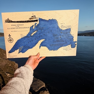 May include: Wooden map of Lake Superior with a blue painted outline, depicting the final voyage of the S.S. Edmund Fitzgerald. The map includes text detailing the ship's route and events. A compass rose and ship illustration are also present.