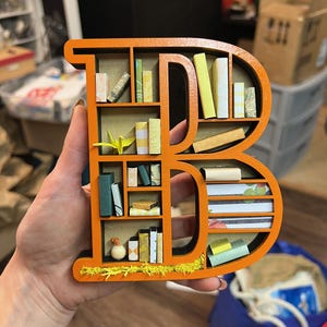 K&ouml;nnte beinhalten: Orangener Holzbuchstabe B in Form eines B&uuml;cherregals mit Miniaturb&uuml;chern und einem kleinen Origami-Vogel im obersten Regal.