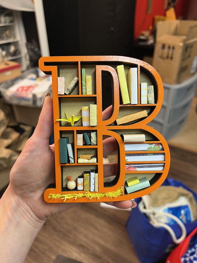 K&ouml;nnte beinhalten: Orangener Holzbuchstabe B in Form eines B&uuml;cherregals mit Miniaturb&uuml;chern und einem kleinen Origami-Vogel im obersten Regal.