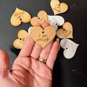 Może przedstawiać: Drewniane ozdoby w kształcie serca z wygrawerowanym napisem „I am enough”. Serca są w różnych odcieniach drewna i bieli, z małym otworem u góry. Ozdoby są rozrzucone na ciemnej powierzchni.