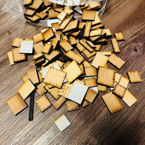 Pode incluir: Uma coleção de pequenas telhas de madeira quadradas de cor castanho claro com bordas mais escuras, espalhadas sobre uma superfície de madeira. Algumas telhas estão em um saco plástico transparente. A imagem inclui algumas telhas retangulares e quadradas.