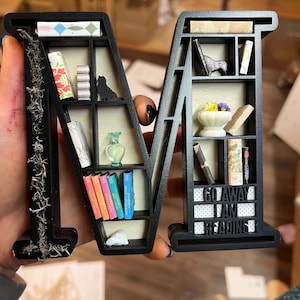 May include: Black wooden letter "M" shaped bookshelf, filled with miniature books, a black cat figurine, a small vase, and the text "GO AWAY I AM READING". The left side of the letter is decorated with dried moss.