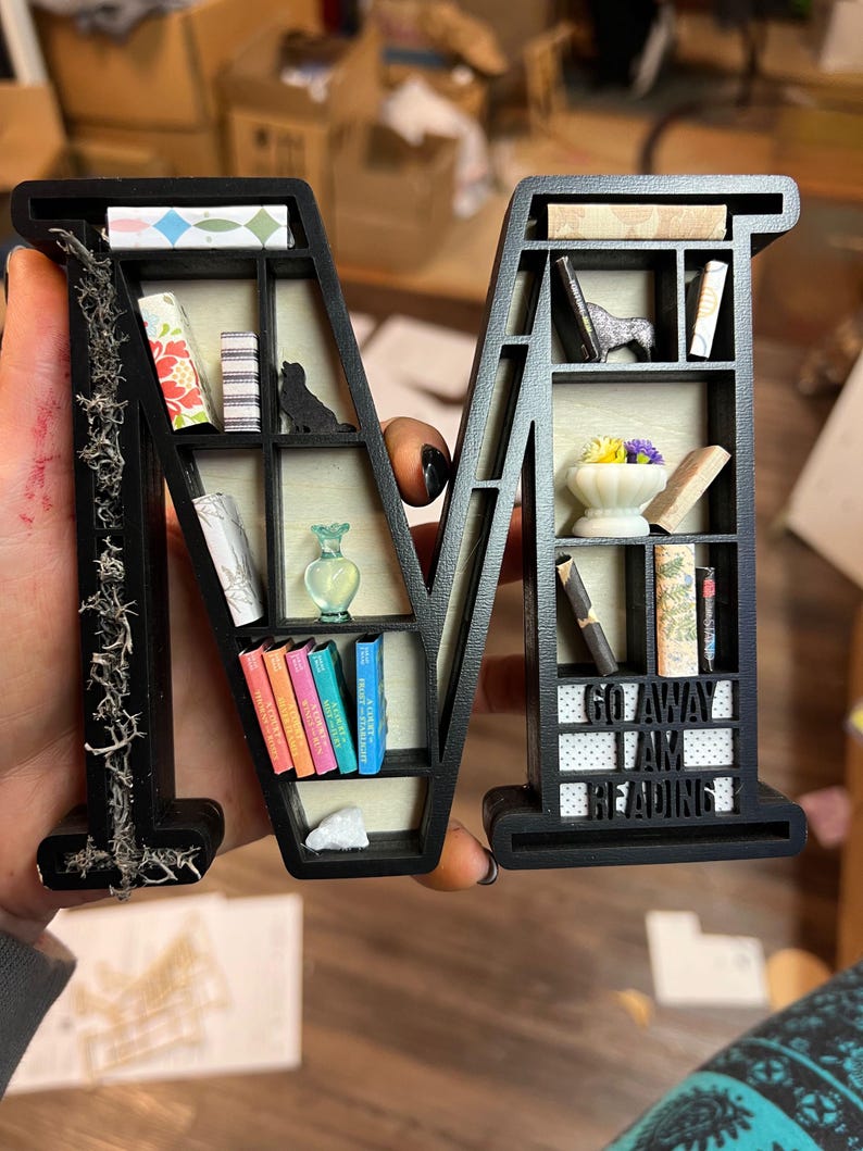 May include: Black wooden letter "M" shaped bookshelf, filled with miniature books, a black cat figurine, a small vase, and the text "GO AWAY I AM READING". The left side of the letter is decorated with dried moss.