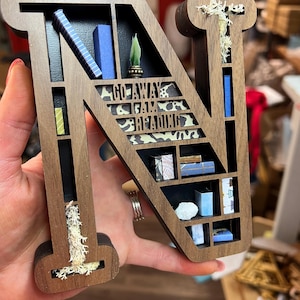 K&ouml;nnte beinhalten: Ein h&ouml;lzerner Buchstabe N, der wie ein B&uuml;cherregal geformt ist, mit Miniaturb&uuml;chern, einer Pflanze und dem Text "Go Away I Am Reading" auf der Vorderseite. Der Buchstabe ist braun mit einer nat&uuml;rlichen Holzoptik und hat ein wei&szlig;es, moosartiges Material in den Spalten.