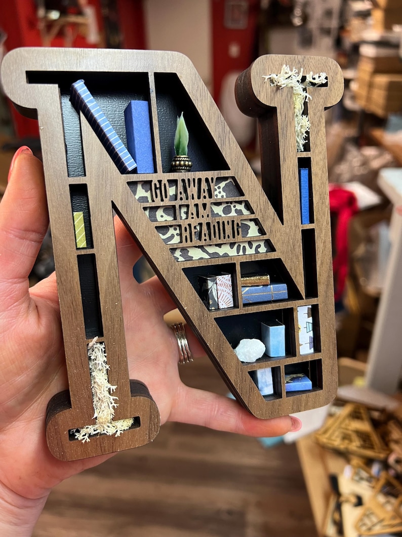 K&ouml;nnte beinhalten: Ein h&ouml;lzerner Buchstabe N, der wie ein B&uuml;cherregal geformt ist, mit Miniaturb&uuml;chern, einer Pflanze und dem Text "Go Away I Am Reading" auf der Vorderseite. Der Buchstabe ist braun mit einer nat&uuml;rlichen Holzoptik und hat ein wei&szlig;es, moosartiges Material in den Spalten.
