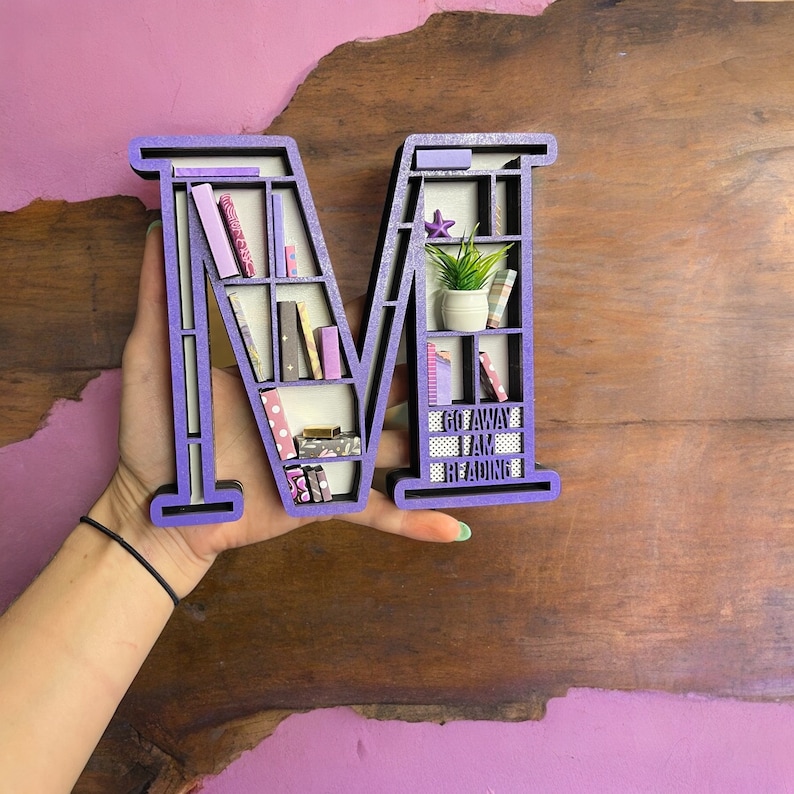 K&ouml;nnte beinhalten: Lilafarbener Holzbuchstabe M in Form eines B&uuml;cherregals mit Miniaturb&uuml;chern, einer Pflanze und einem Schild mit der Aufschrift "Go Away I Am Reading".