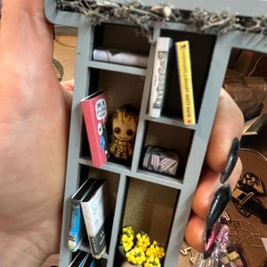 May include: Miniature gray bookshelf with several tiny books, a Groot figurine, and a small vase of yellow flowers. The bookshelf is decorated with faux greenery along the top. The books have various colors and titles.