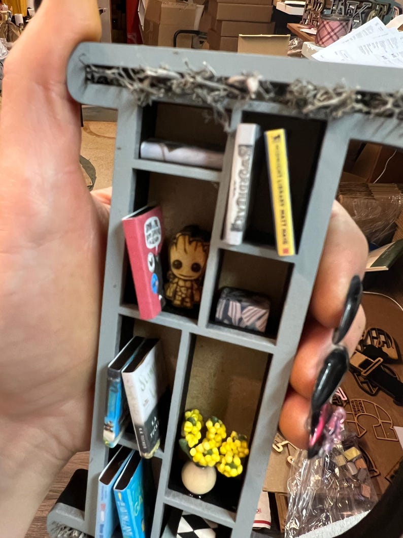 May include: Miniature gray bookshelf with several tiny books, a Groot figurine, and a small vase of yellow flowers. The bookshelf is decorated with faux greenery along the top. The books have various colors and titles.