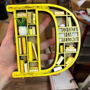 K&ouml;nnte beinhalten: Ein gelber Holzbuchstabe D in Form eines B&uuml;cherregals mit Miniaturb&uuml;chern und einer kleinen Topfpflanze. Das B&uuml;cherregal tr&auml;gt den Text "Go Away I Am Reading" auf der Vorderseite.