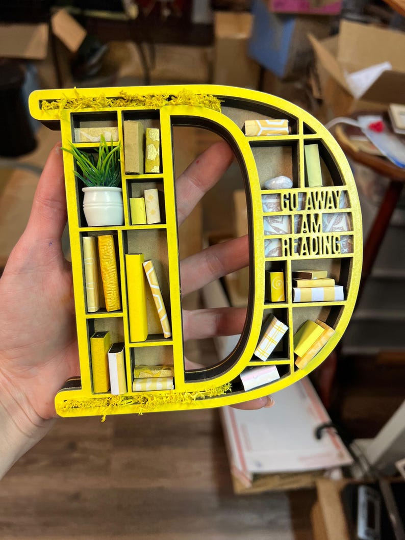 K&ouml;nnte beinhalten: Ein gelber Holzbuchstabe D in Form eines B&uuml;cherregals mit Miniaturb&uuml;chern und einer kleinen Topfpflanze. Das B&uuml;cherregal tr&auml;gt den Text "Go Away I Am Reading" auf der Vorderseite.