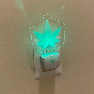 May include: A white night light with a green cannabis leaf design. The night light plugs into a standard wall outlet and emits a soft green glow. The leaf is the focal point of the design.
