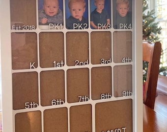 May include: White framed photo display with spaces for school year photos. Includes spaces for PK1 through 12th grade, plus the name "ROBERT" and the year "Est 2018". The display has a clean, modern design.