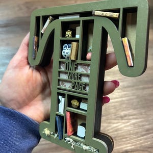 May include: A green wooden letter "T" shaped bookshelf with miniature books, a cupcake, and an owl figurine. The text "ONE MORE PAGE" is visible. The bookshelf is approximately 8 inches tall.