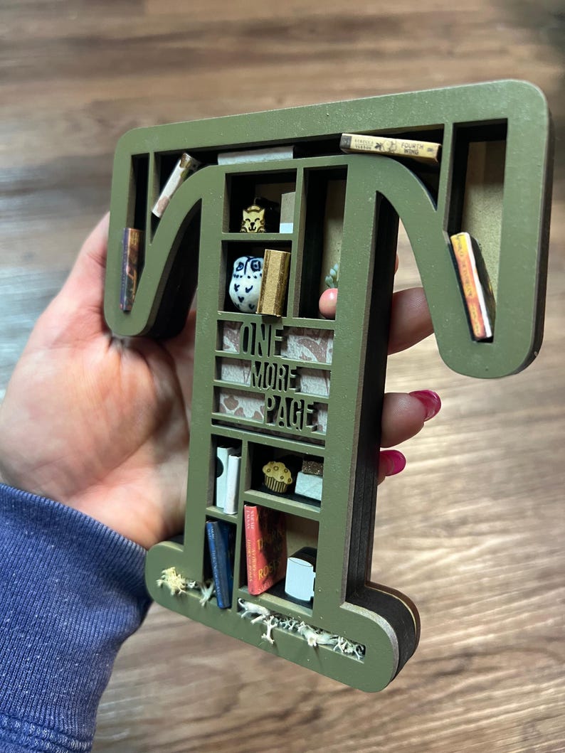 May include: A green wooden letter "T" shaped bookshelf with miniature books, a cupcake, and an owl figurine. The text "ONE MORE PAGE" is visible. The bookshelf is approximately 8 inches tall.