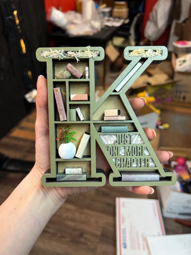 K&ouml;nnte beinhalten: Ein gr&uuml;ner Holzbuchstabe K in Form eines B&uuml;cherregals mit Miniaturb&uuml;chern, einer Pflanze und einer wei&szlig;en Vase. Der Buchstabe tr&auml;gt den Text "Just One More Chapter" an der Seite.
