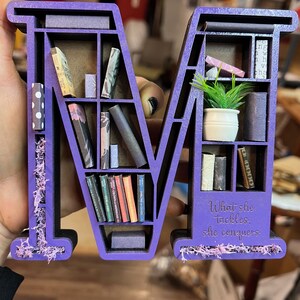May include: A purple wooden letter "M" shaped bookshelf, filled with miniature books and a small potted plant. The base of the shelf has the quote "What she tackles, she conquers". Decorative moss accents the sides.