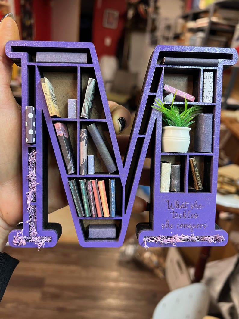 May include: A purple wooden letter "M" shaped bookshelf, filled with miniature books and a small potted plant. The base of the shelf has the quote "What she tackles, she conquers". Decorative moss accents the sides.