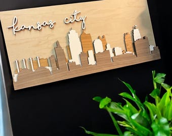 May include: Wooden wall art featuring the Kansas City skyline. The artwork is made of layered wood in shades of brown and beige, with the city's name in cursive script. The piece is mounted on a black wall, with a green plant in the foreground.