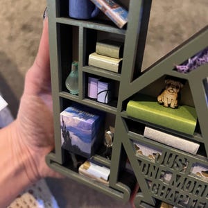 May include: A green wooden shelf in the shape of the letter "K" filled with miniature books, a blue mug, a glass bottle, and a pug figurine. The text "JUST ONE MORE CHAPTER" is visible.