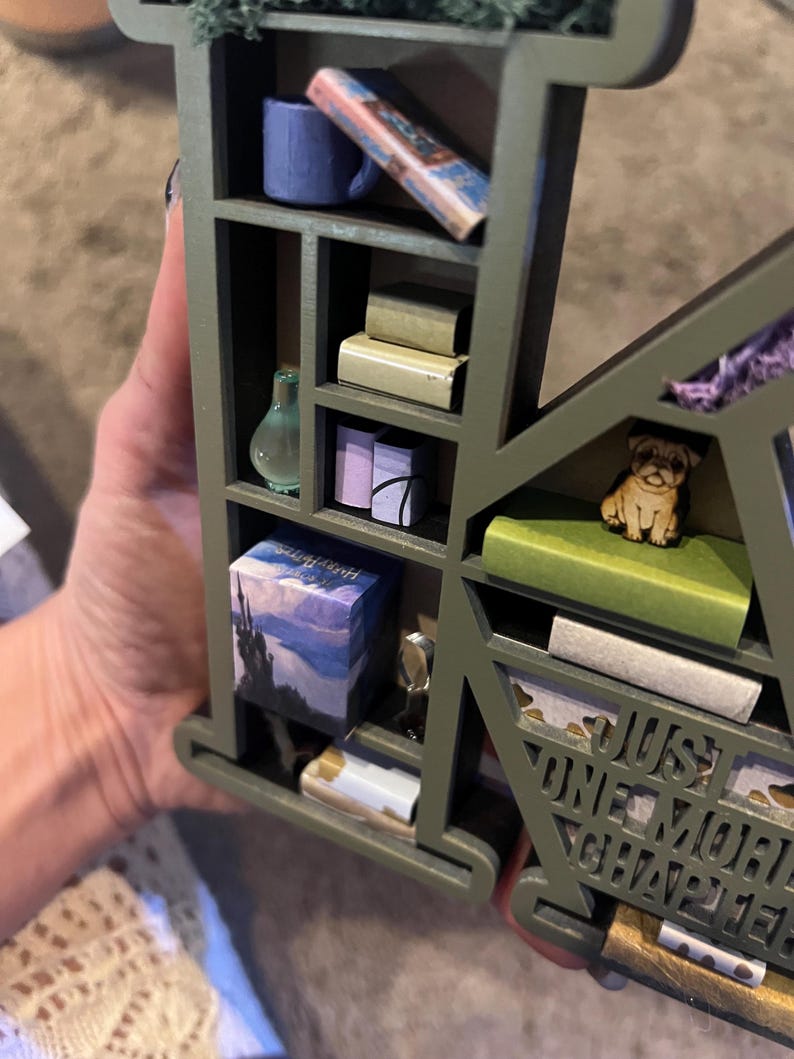 May include: A green wooden shelf in the shape of the letter "K" filled with miniature books, a blue mug, a glass bottle, and a pug figurine. The text "JUST ONE MORE CHAPTER" is visible.
